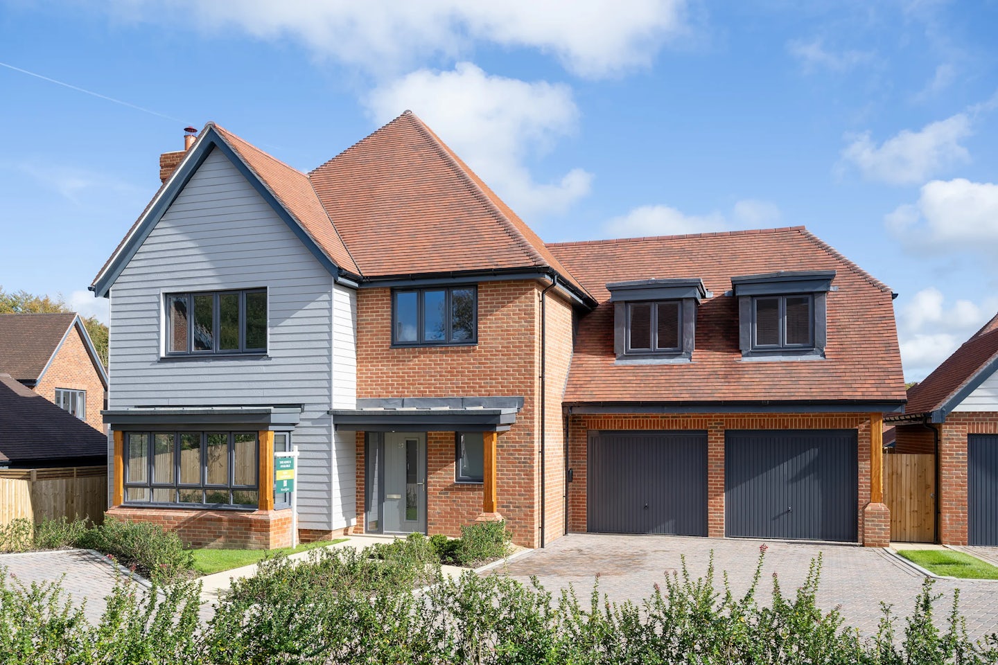 A modern two-story house with a sloped red-shingled roof and a mixed brick and siding exterior. The facade features a prominent bay window on the left, two dormer windows above a double garage on the right, and a modest covered porch at the front entrance. A light brick driveway leads up to the house, bordered by neatly trimmed shrubs and greenery. The setting is bright and sunny under a mostly clear sky.