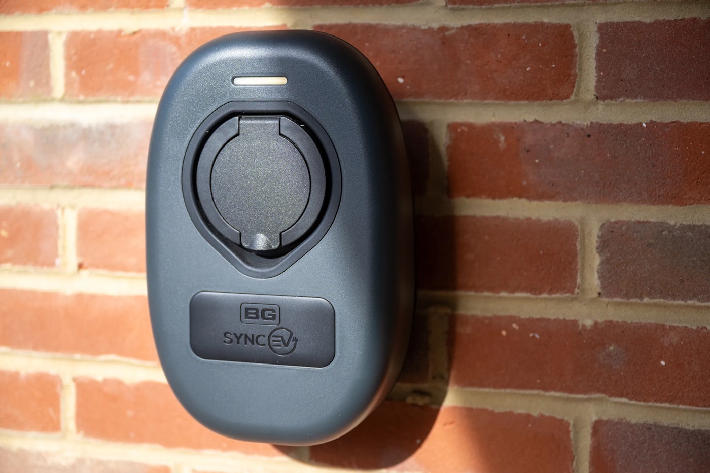 A grey “BG Sync EV” electric vehicle charging point mounted on a reddish-brown brick wall, featuring a round socket cover and subtle branding, representing accessible, eco-conscious transport infrastructure in a residential or commercial setting.
