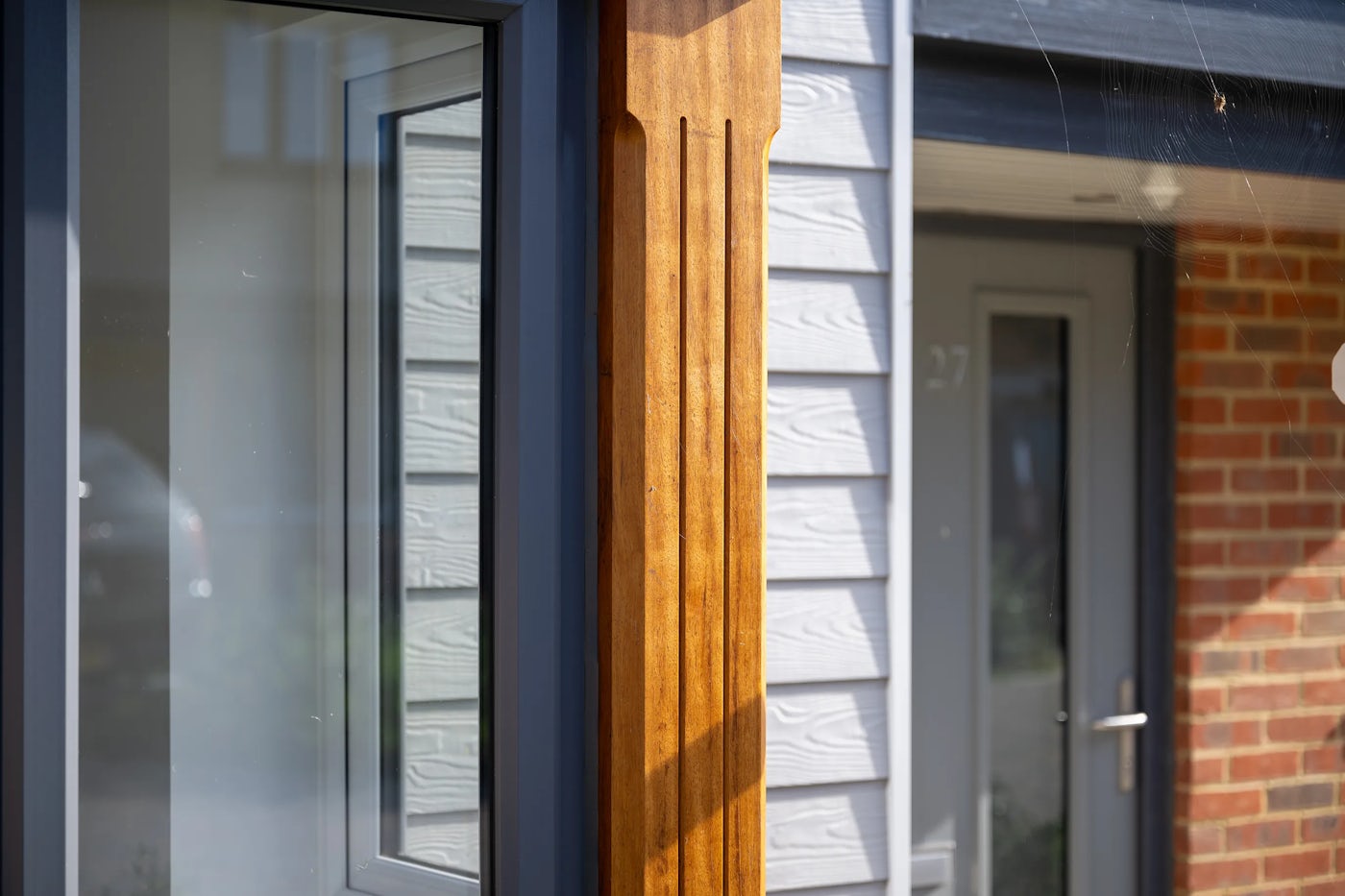 A close-up of a modern residential entrance, featuring a vertically grooved wooden column beside a large reflective window. In the background, a white door marked with the number “27” is partially visible, framed by white siding and a section of reddish brick wall.