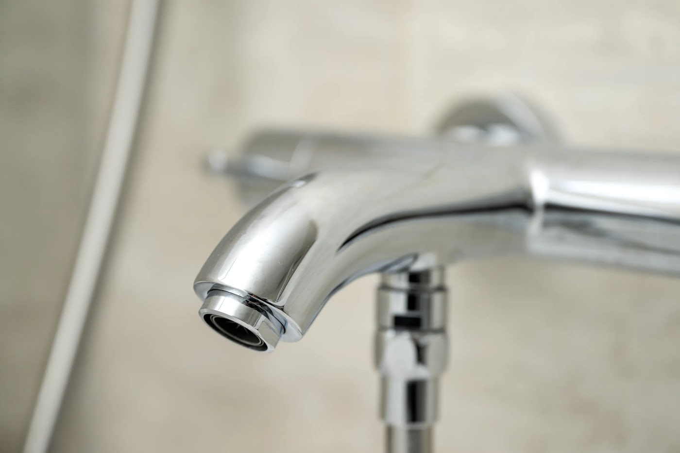 Close-up of a modern chrome bathroom faucet with a sleek, reflective finish, set against a pale, neutral-toned tiled wall.