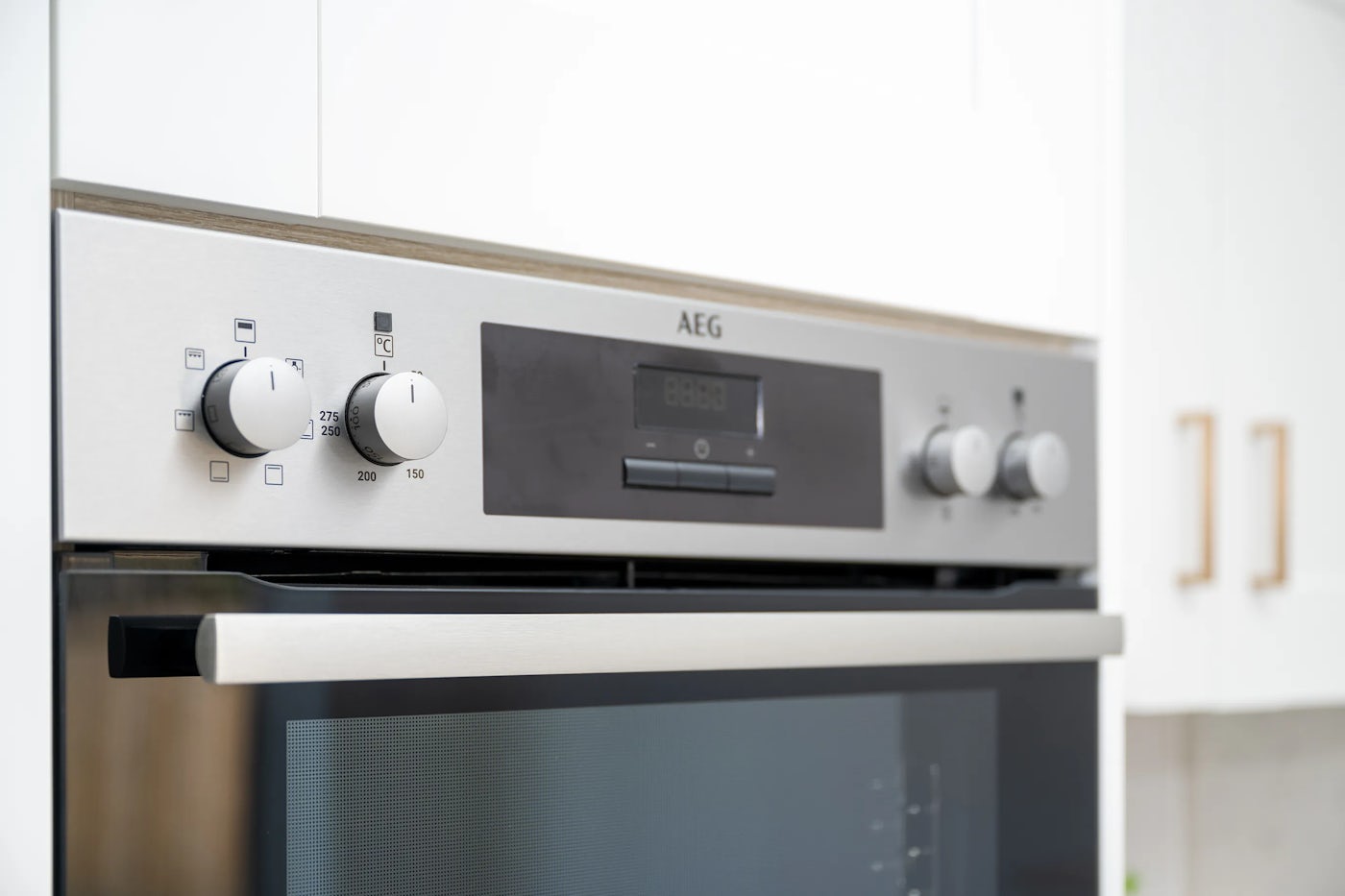 Close-up of a stainless steel AEG oven control panel featuring four round knobs and a central digital display. The left knobs control oven function and temperature, with temperature markings from 50°C to 250°C. The digital display, flanked by small buttons, shows additional settings. The right knobs likely adjust time and auxiliary functions. A horizontal handle and glass window are visible on the oven door below.