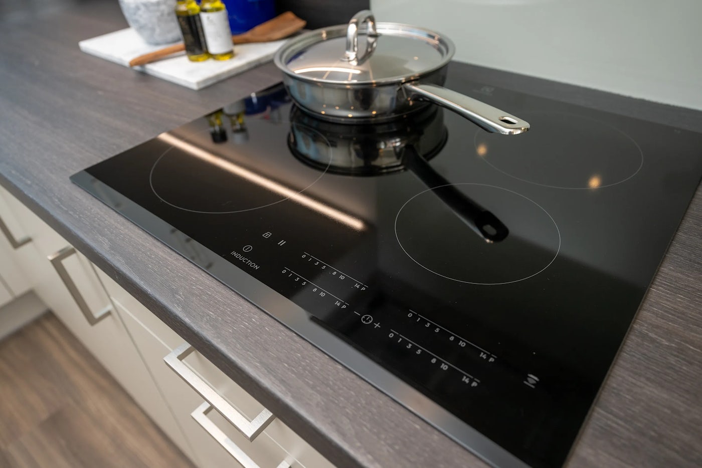 A modern kitchen countertop featuring a sleek black induction cooktop with illuminated touch controls. A stainless steel pan with a lid rests on one of the active cooking zones. In the background, olive oil bottles and a wooden cutting board add warmth and functionality to the contemporary setting.