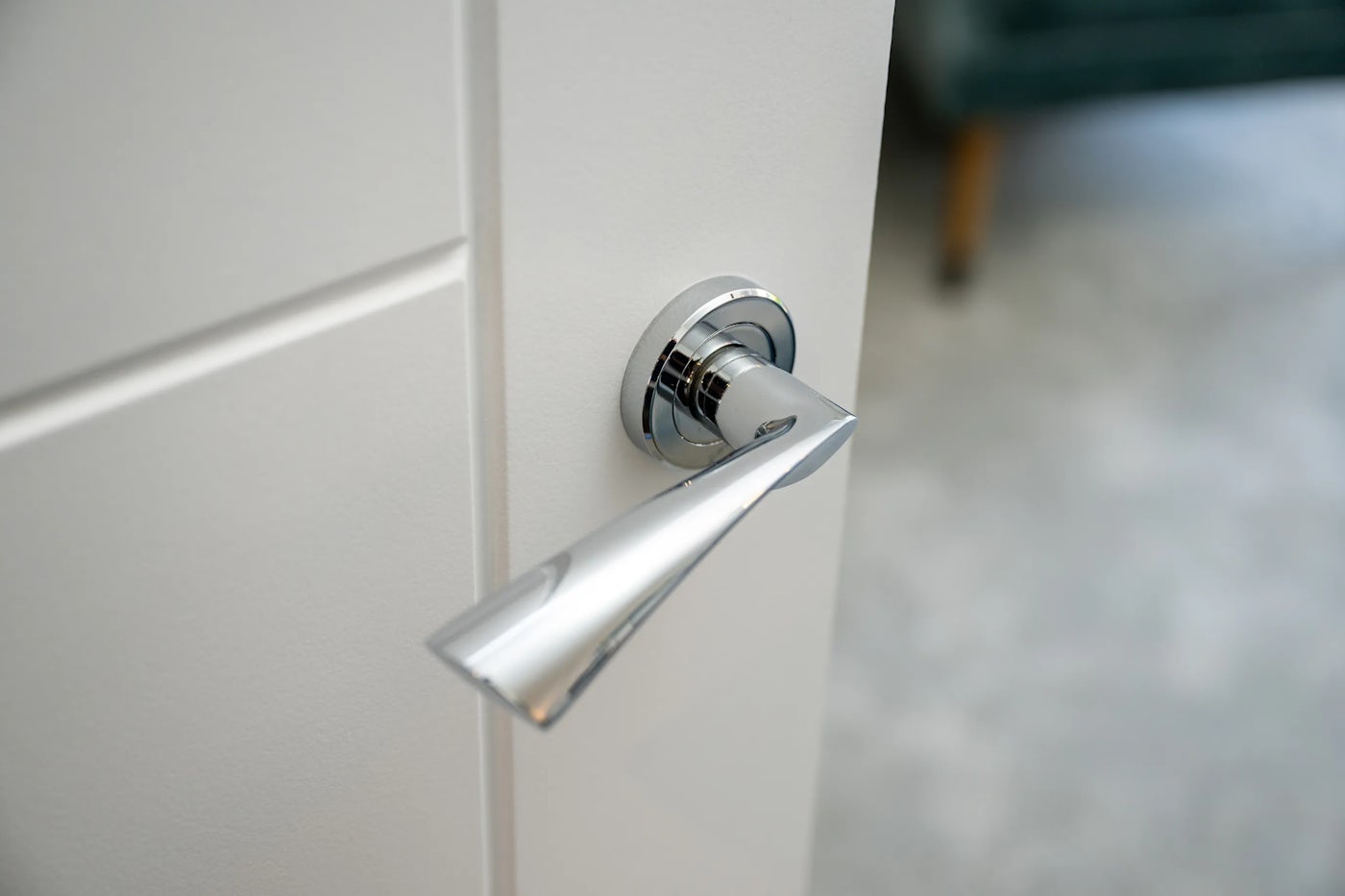 Close-up of a sleek, metallic door handle mounted on a white door, with a soft-focus view of a carpeted interior in the background, including the leg of a nearby chair or sofa