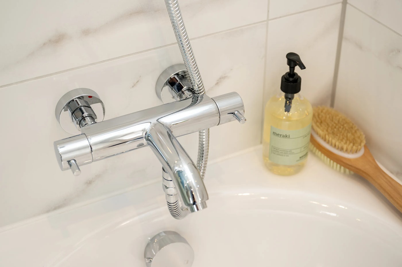 A bathroom sink area featuring a chrome faucet with hot and cold handles and an attached showerhead on a flexible metal hose. Beside the faucet sits a pump bottle labeled “meraki” and a wooden bristle brush. The background consists of white tiles with subtle grey veining.