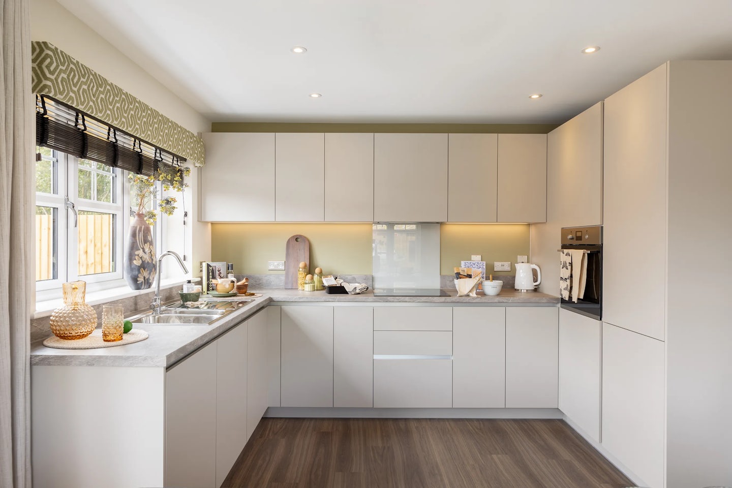 Modern kitchen with light cabinets, marble counters, and wooden flooring, featuring a double sink under a window with a patterned valance, surrounded by small appliances, decorative jars, and warm under-cabinet lighting.