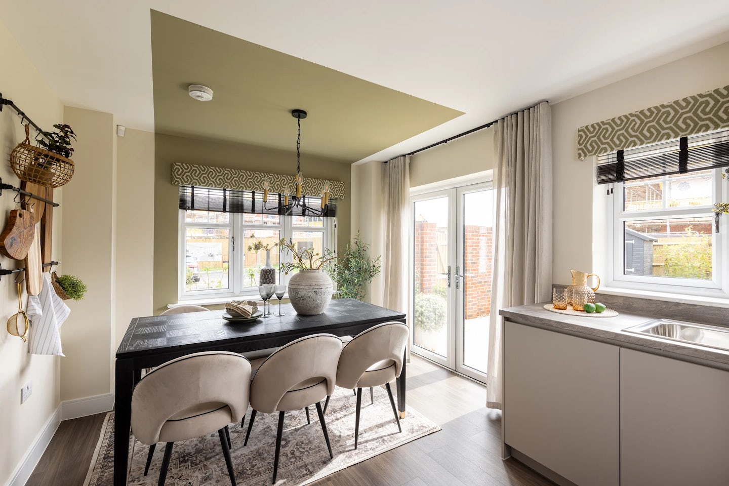 Modern dining area with a black rectangular table and four grey upholstered chairs, set beneath a hanging light fixture. A large decorative vase with green foliage sits at the centre of the table. The space adjoins a sleek kitchen with a countertop displaying a bowl of lemons, cutting board, and pitcher. Floor-to-ceiling windows with patterned valances overlook a garden, and a green ceiling visually defines the dining zone within the open-plan layout.