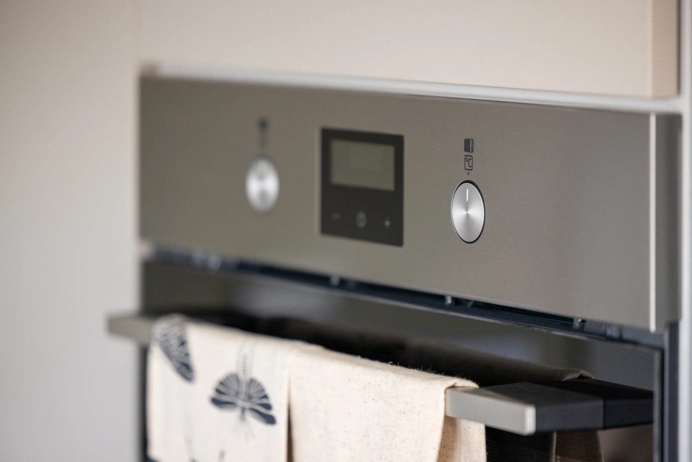 Close-up of a built-in kitchen oven’s control panel, featuring two round silver knobs—one on each side of a central digital display. A white towel hangs neatly from the oven door handle below, suggesting a tidy, functional cooking space.