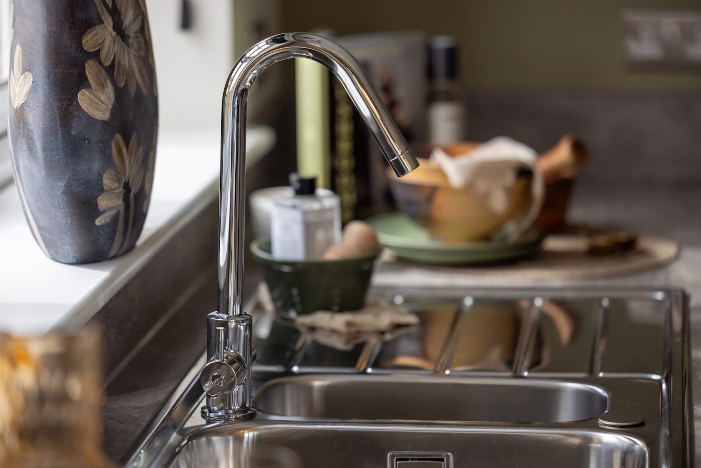 A close-up view of a modern kitchen sink area featuring a sleek, chrome-finished faucet with a curved spout positioned above a single-basin stainless steel sink. To the right of the sink is a draining board. Behind the sink, the dark countertop holds a decorative ceramic vase with floral patterns, a green bowl, a bottle of liquid soap, and a wooden bowl with a folded cloth inside. Soft natural light enters from the left, adding warmth to the clean, contemporary setup.