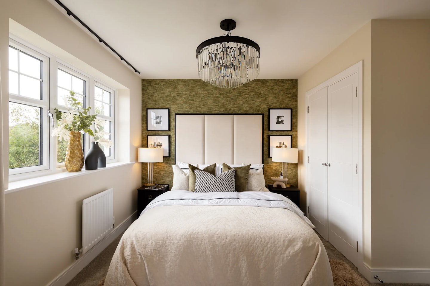 A softly lit bedroom styled with elegance and warmth. A centrally placed bed features layered white and beige bedding, framed by twin bedside tables with matching lamps. Behind the bed, a textured green wallpaper acts as a backdrop, adorned with two symmetrical framed artworks. A chandelier adds a touch of refinement from above. To the left, natural light filters in through a window dressed with a vase of fresh flowers, while to the right, a crisp white double-door closet balances the space. The room feels cosy yet refined, blending decorative charm with functional comfort.