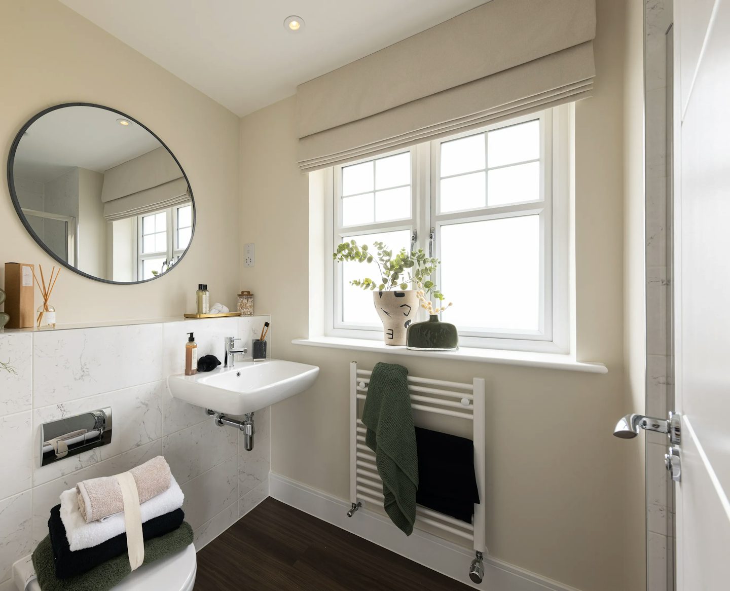 Modern bathroom with a minimalist design, featuring a round mirror above a white sink, a window with beige blinds, and a radiator draped with folded towels. A shelf above the toilet holds toiletries including a soap dispenser, reed diffuser, and small container. A green plant in a vase sits on the window sill, and the space is styled with neutral tones and a wooden floor..