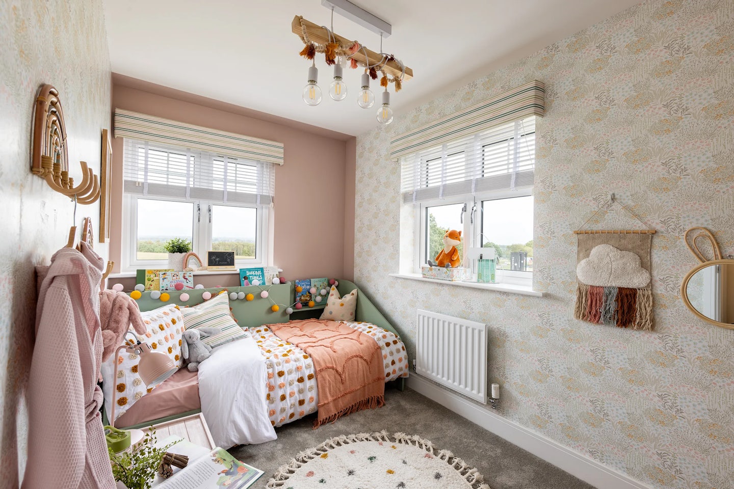 A softly lit children’s bedroom with a playful, coordinated decor. A single bed dressed in colourful polka dot and striped bedding is set against light pink walls, some adorned with patterned wallpaper. Two windows with white blinds and patterned curtains allow natural light into the space. To the left, a wooden coat rack displays a pink robe and a stuffed toy, while the right wall features a small round mirror and a decorative cloud-shaped wall hanging with tassels. A circular rug with a star motif rests on the wooden floor, and a ceiling light fixture with exposed bulbs adds a warm glow.