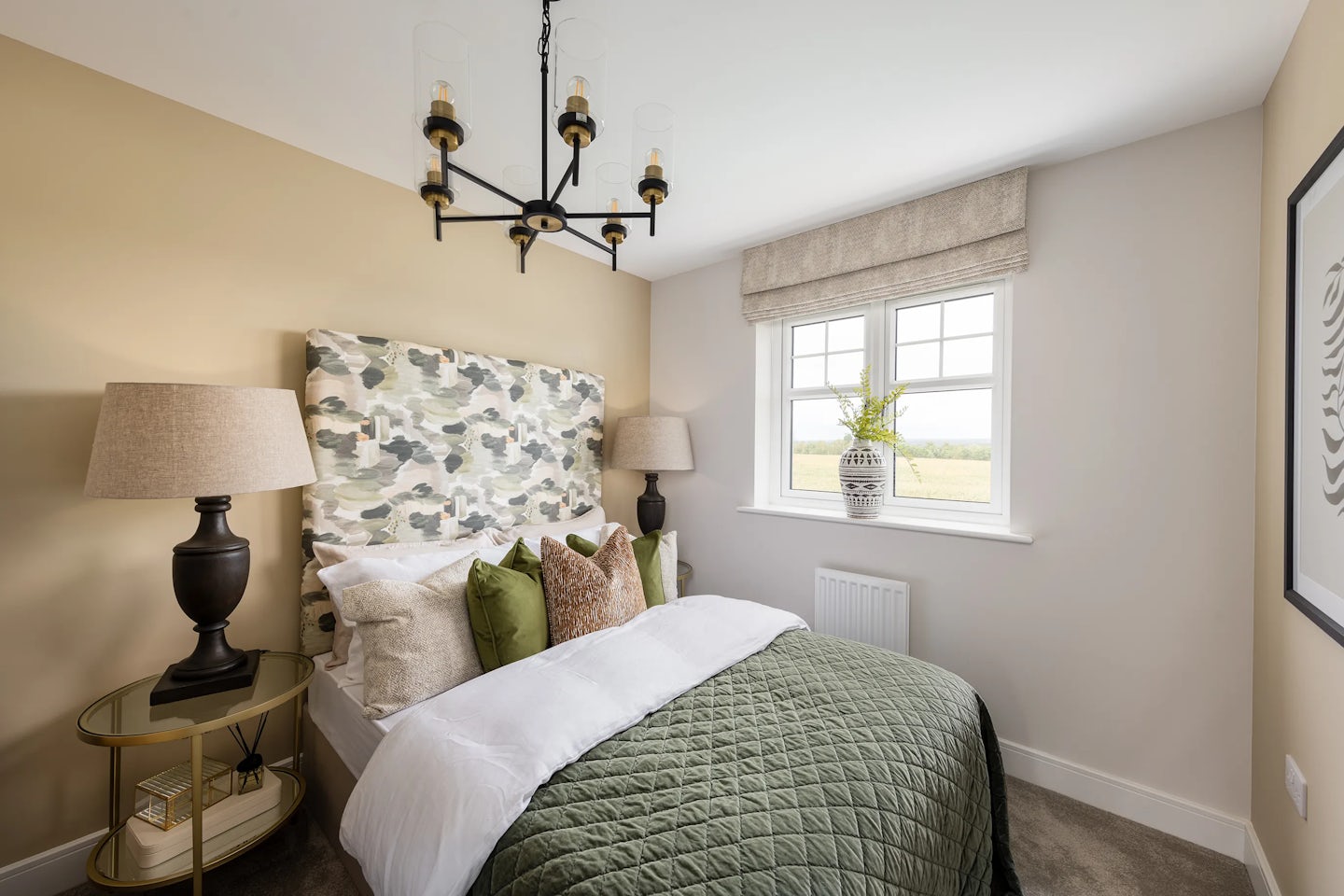 A softly lit bedroom with a green and beige colour palette. The bed features a camouflage-patterned headboard, green quilted bedspread, and coordinating pillows. To the left, a bedside table holds a classic lamp and decorative items. A five-light chandelier hangs overhead, while a window with a beige roman shade lets in natural light. A vase with greenery sits on the windowsill, complementing the neutral-toned walls and framed artwork on the right. The space feels calm, cohesive, and thoughtfully styled.
