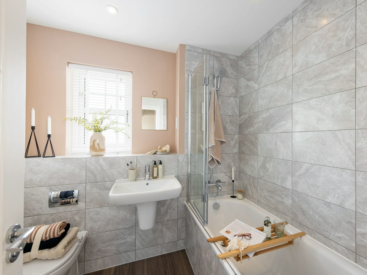 A modern bathroom featuring a grey-tiled wall and wooden floor, with a bathtub on the left fitted with a glass shower screen and a wooden bath tray holding a book, candle, and bottle. A sink with assorted toiletries sits beneath a mirror, while a toilet is positioned nearby. The window has white blinds and a sill adorned with candles and a vase of flowers. A towel hangs beside the tub, and the space is brightly lit, evoking a clean, serene atmosphere.