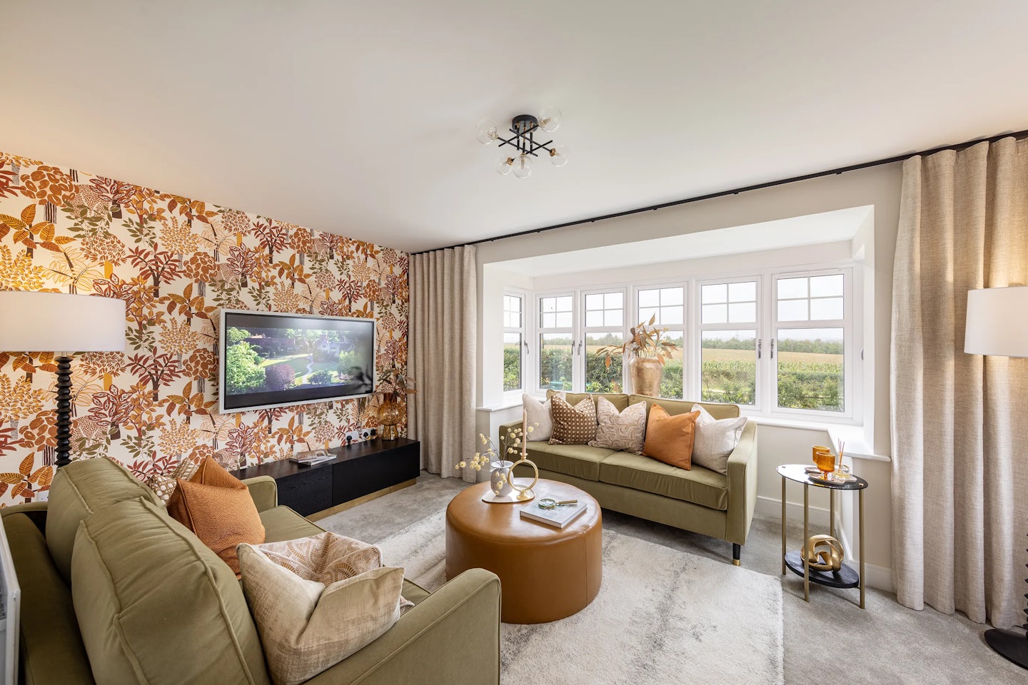 A contemporary living room with a cozy, curated atmosphere. A green and a beige sofa face a round brown ottoman, doubling as a coffee table. Behind the green sofa, a bold floral wallpaper in orange, brown, and white adds vibrancy, complemented by a mounted TV displaying a tranquil nature scene. Tall lamps with white shades flank a large window overlooking a green field, bringing in soft natural light. Decorative accents on a round side table and a modern ceiling light complete the inviting, design-conscious space.