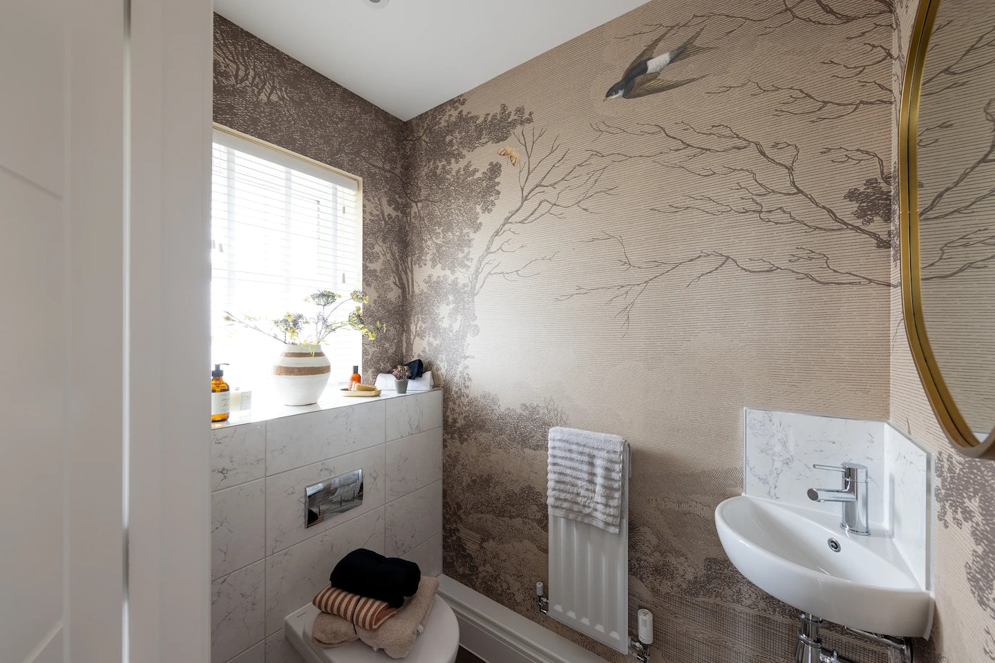 A compact, stylish bathroom with natural light filtering through slatted blinds. A feature wall with botanical wallpaper—showing tree branches and a small bird—adds visual charm. Below the window, a shelf holds a potted plant, toiletries, and soap dispensers. A towel rests on the closed toilet lid, while another hangs from a white radiator. A petite sink and mirror complete the space, creating a calm, modern atmosphere.