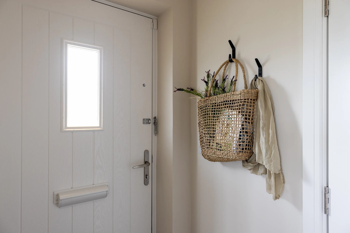 Interior view of an entryway featuring a white door with a rectangular window near the top and a silver mail slot below. To the right, a pair of black wall hooks hold a woven basket of greenery and a pale scarf, adding a welcoming, decorative touch.