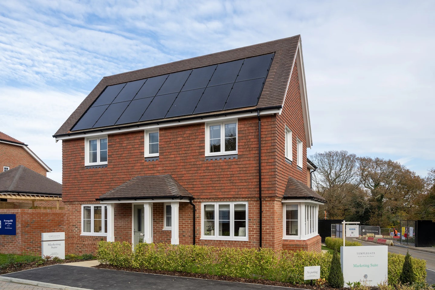 Two-story show home with rooftop solar panels, red brick exterior, and white-framed windows, surrounded by a small landscaped front garden. Signs in front promote 'Temple Park' development. Another similar home and leafy trees appear in the suburban background beneath a partly cloudy sky.