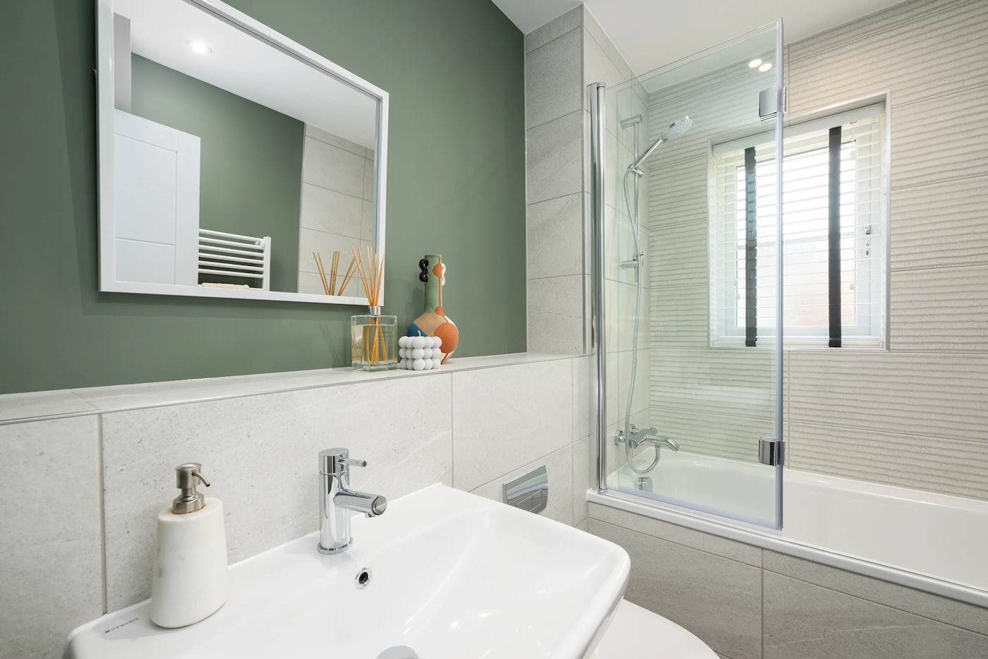 A modern bathroom with a clean, minimalist design. A white rectangular sink with a chrome faucet sits beneath a large mirror, accompanied by a soap dispenser and decorative items including a reed diffuser and a small container of cotton balls. To the right, a bathtub with a glass shower screen and showerhead is set against a textured wall. A window with white blinds above the tub lets in natural light. The room features green and beige tiles, creating a calm, spa-like atmosphere.