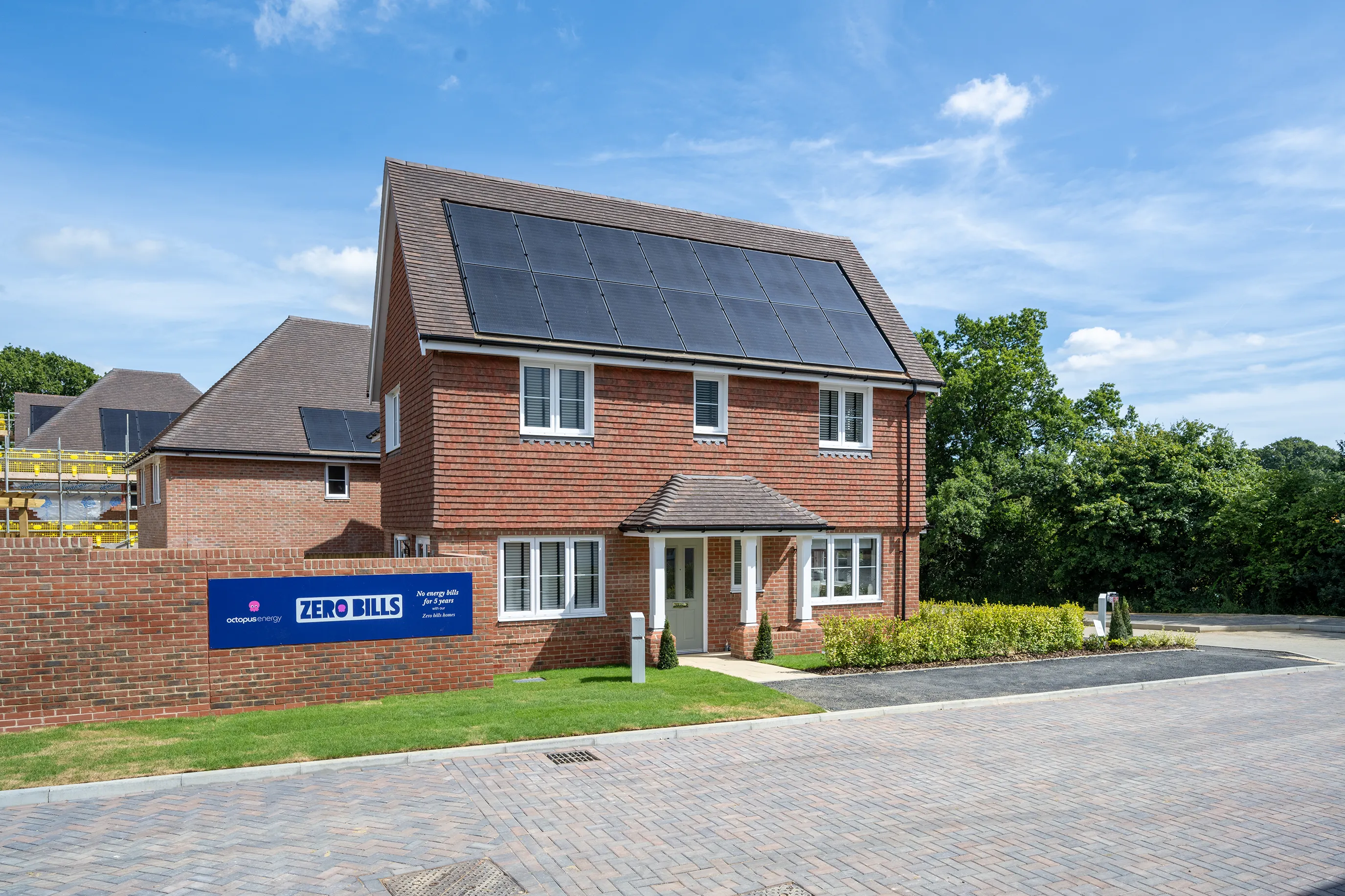 A modern two-storey brick house with white-framed windows, a small front porch, and rooftop solar panels. A sign in front reads “ZERO BILLS” with logos for Octopus Energy and ilke Homes, promoting “all energy bills paid for 10 years” and “The UK’s first zero bills home.” Construction activity is visible in the background, indicating a new housing development.