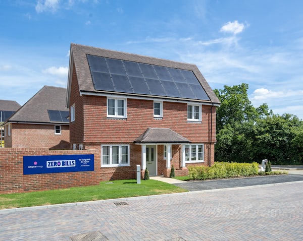 A modern two-storey brick house with white-framed windows, a small front porch, and rooftop solar panels. A sign in front reads “ZERO BILLS” with logos for Octopus Energy and ilke Homes, promoting “all energy bills paid for 10 years” and “The UK’s first zero bills home.” Construction activity is visible in the background, indicating a new housing development.