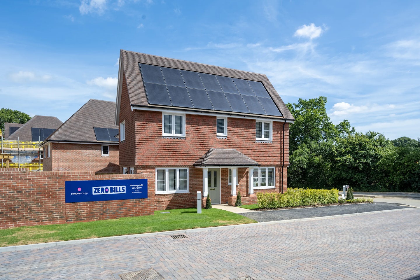 A modern two-storey brick house with white-framed windows, a small front porch, and rooftop solar panels. A sign in front reads “ZERO BILLS” with logos for Octopus Energy and ilke Homes, promoting “all energy bills paid for 10 years” and “The UK’s first zero bills home.” Construction activity is visible in the background, indicating a new housing development.