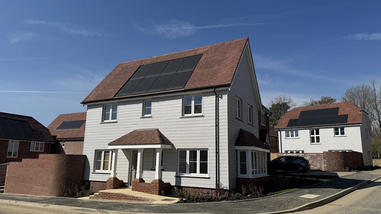 Modern residential street featuring detached homes with pitched roofs fitted with solar panels. The scene is bathed in sunlight under a clear blue sky, suggesting a warm, energy-efficient neighbourhood prioritising renewable resources.