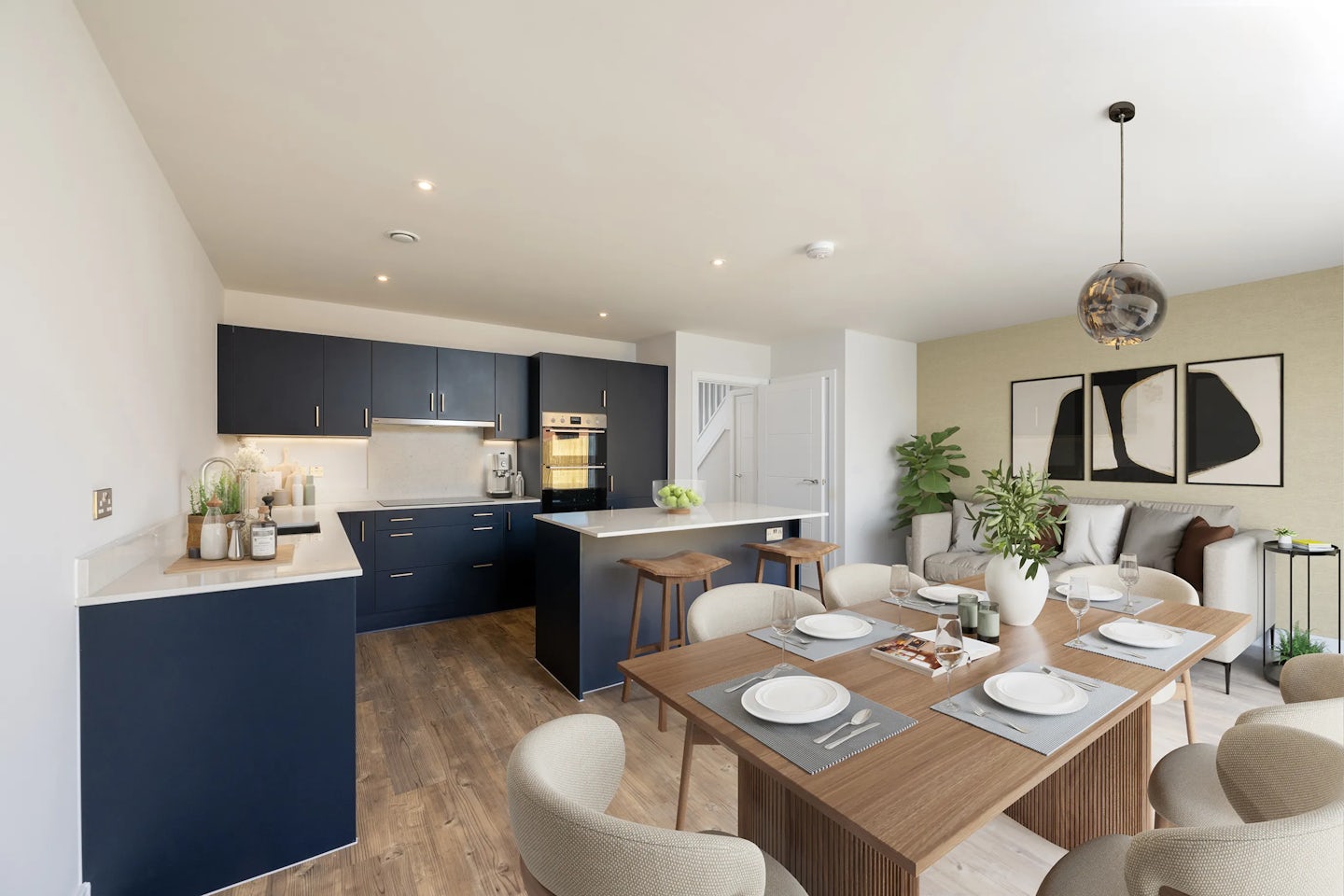 A contemporary open-plan kitchen and dining area featuring dark blue cabinets with gold handles, a central island with two wooden bar stools, and a wooden dining table set for six. The space is styled with white countertops, cushioned dining chairs, and warm lighting from recessed and pendant fixtures. Decorative plants and curated accents introduce greenery and personality, while a relaxed living area with a sofa and wall art sits in the background.