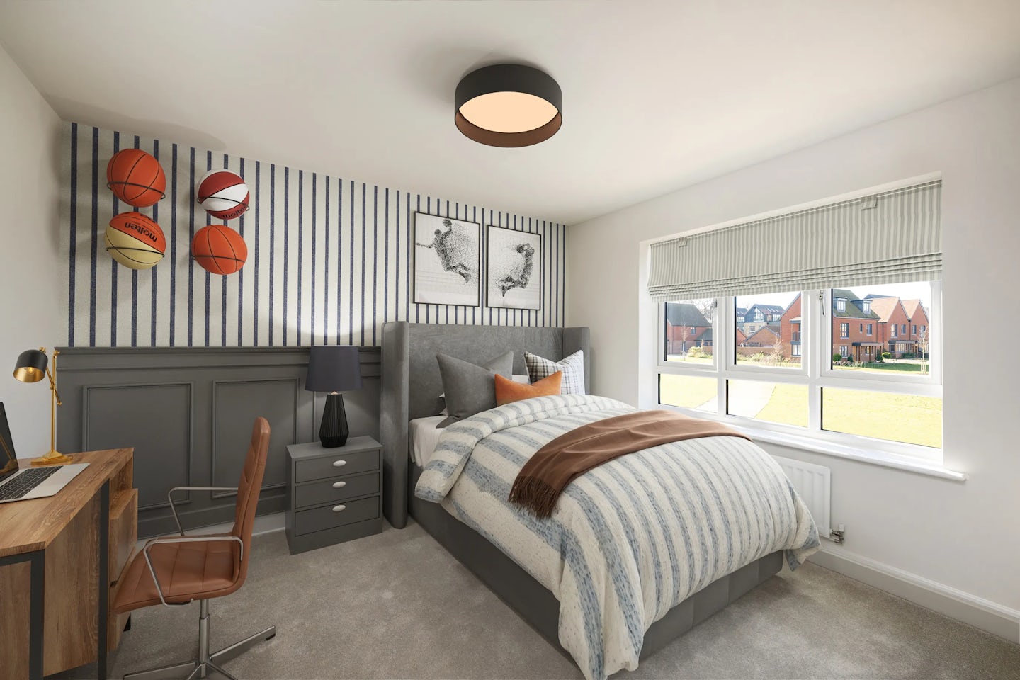 A modern bedroom with a bed featuring a striped duvet and brown throw, set beside a nightstand with a lamp. A desk with a laptop and chair sits under a window overlooking suburban houses and lawn. Decorative basketballs and framed art hang on the wall, creating a sporty, personalised atmosphere.