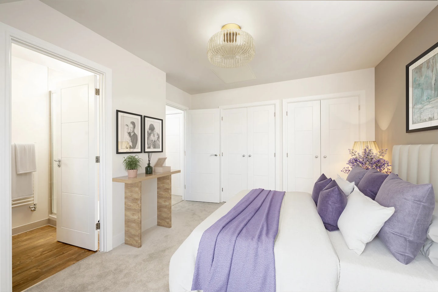 A spacious, modern bedroom featuring a neatly made bed with white linens, accented by purple cushions and a folded purple throw. A nightstand beside the bed holds a lamp and a vase of purple flowers. Across the room, a wooden console table displays a small plant and sits beneath two framed artworks. Multiple white panelled doors and a sculptural ceiling light complete the room’s elegant, serene aesthetic.