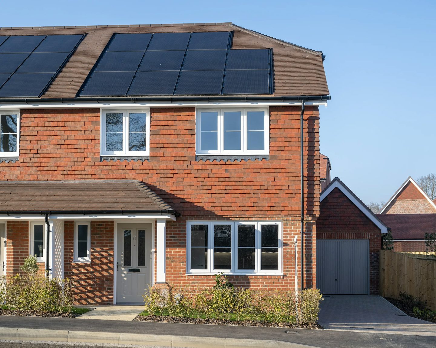 Modern two-storey brick house with white-framed windows, solar panels on the roof, and an attached garage on the right. A paved driveway leads past a small front garden with neatly trimmed shrubs.
