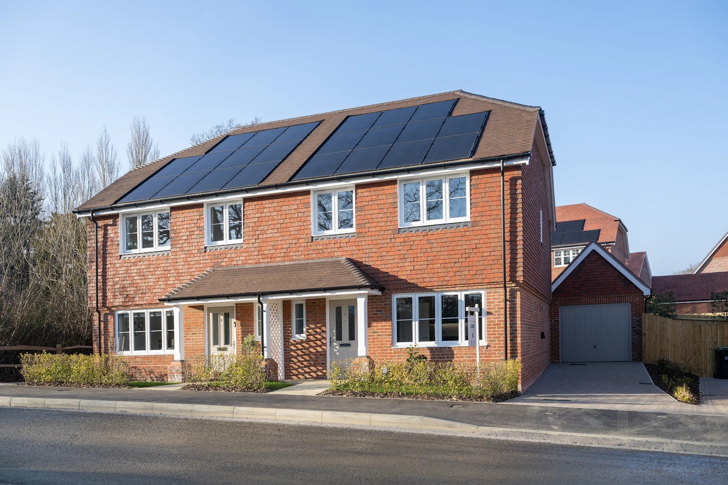 Semi-detached brick house with solar panels on the roof, white-framed windows, and two front doors, indicating dual occupancy; a paved driveway leads to a right-side garage, set in a suburban neighbourhood with clear skies and distant trees.