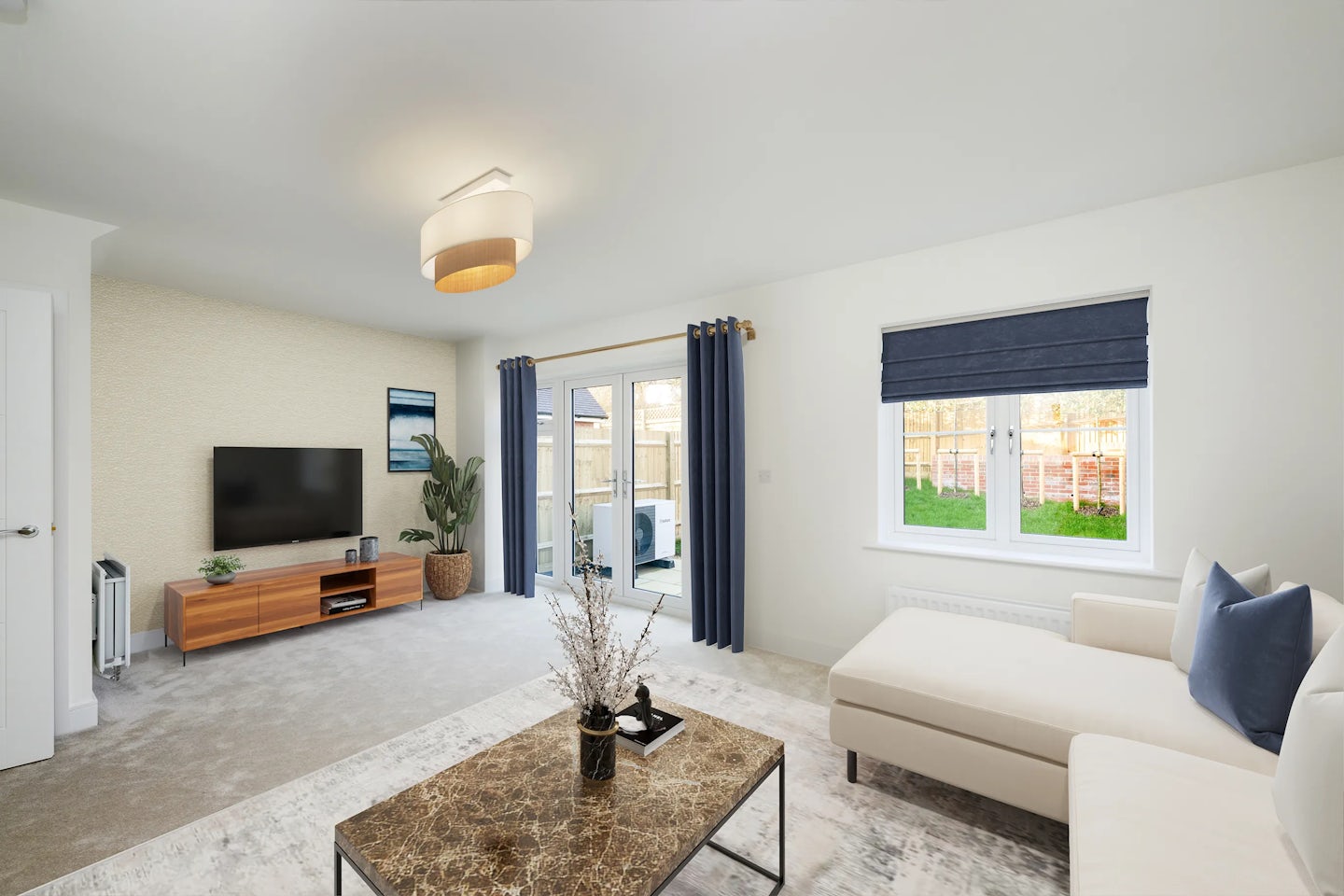 A modern living room with a spacious beige sectional sofa accented by blue throw pillows, facing a flat-screen TV on a wooden stand. A round marble coffee table with decorative objects sits in the centre, while a tall potted plant and a framed picture add character to the corner. Natural light fills the room through a large window with blue blinds and a sliding glass door draped in matching curtains, opening to a green lawn enclosed by a wooden fence. A sculptural ceiling light completes the bright, minimalist space.