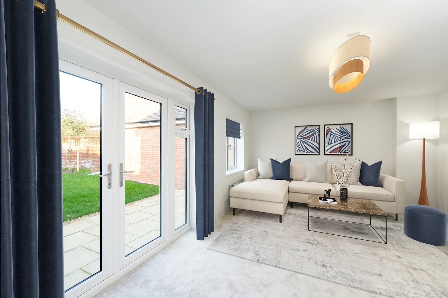 A modern living room with a large glass door opening onto a patio and garden, furnished with a light-toned sectional sofa, a marble coffee table, and a blue ottoman. Two framed abstract artworks hang above the sofa, flanked by dark blue curtains. A contemporary ceiling light and a floor lamp create a warm, inviting atmosphere.