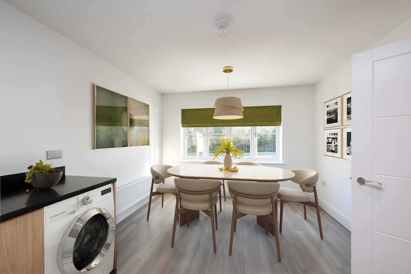 A light-filled dining space with a white rectangular table set for six, each chair featuring a black seat and backrest. A vase of bright yellow flowers acts as the table’s cheerful centrepiece. Above, a minimalist pendant light hangs from the ceiling. To the left, a worktop holds a bowl of grapes, with a washing machine tucked underneath. The backdrop includes green window blinds, framed artwork, and photographs adorning the white walls, all grounded by pale wood flooring that enhances the room’s clean, contemporary feel.