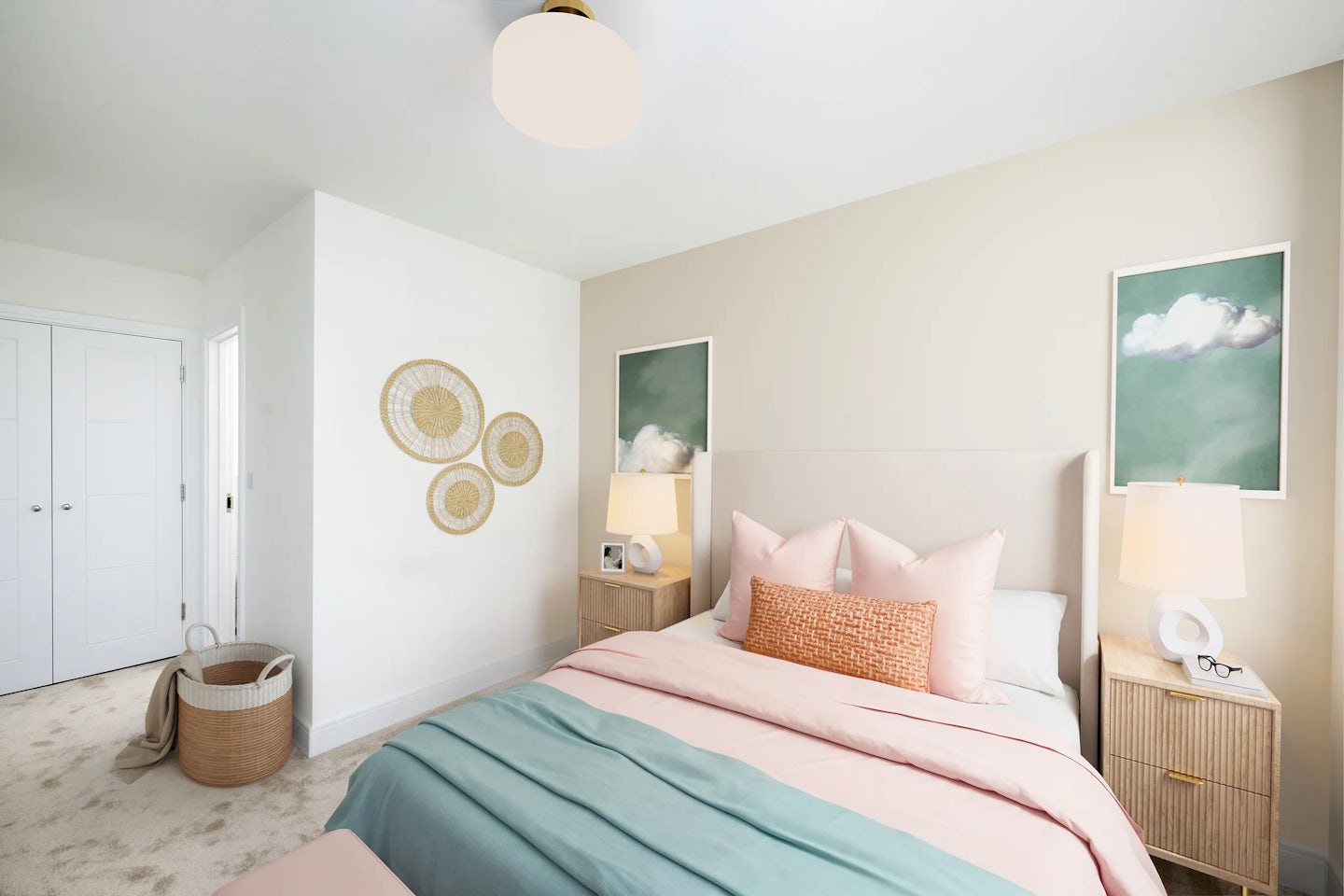 A softly lit, modern bedroom with a minimalist design. A bed with light pink and teal bedding, layered with pink and orange cushions, sits centrally against a pale green wall. Above the bed hang two framed cloud illustrations. Wooden nightstands on either side each hold a lamp and decorative accents. To the left, three woven circular wall hangings add texture, while a wicker basket rests near double closet doors. The space feels calm, airy, and thoughtfully styled.