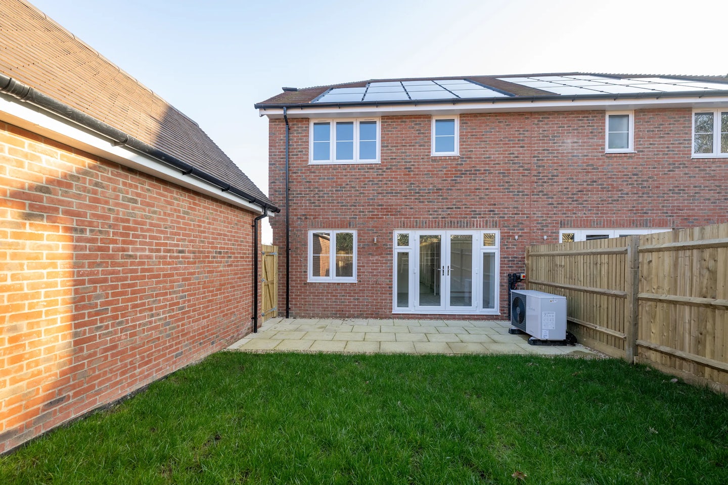 Backyard of a brick house featuring a small patio with light-coloured tiles and a neatly trimmed lawn. The house roof is fitted with solar panels, and a glass door opens to the patio from a room with large windows. An air conditioning unit is positioned beside a wooden fence on the right.