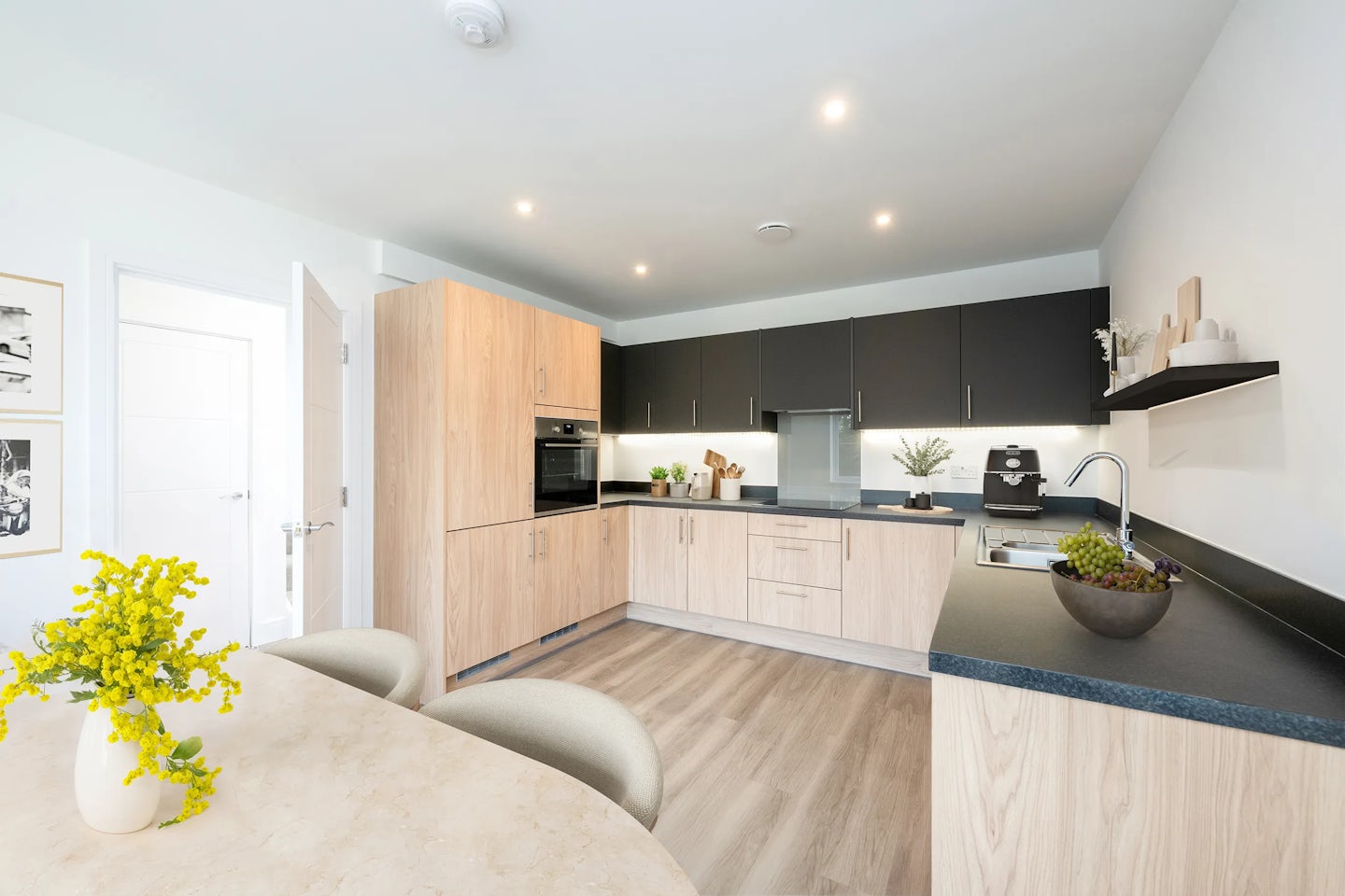 Modern kitchen with light wood cabinetry and black countertops, featuring built-in appliances, a stainless steel sink, and a coffee machine. Decorative plants and bowls add warmth to the clean, contemporary design. Light wood flooring and a dining table with chairs complete the open, well-lit space.