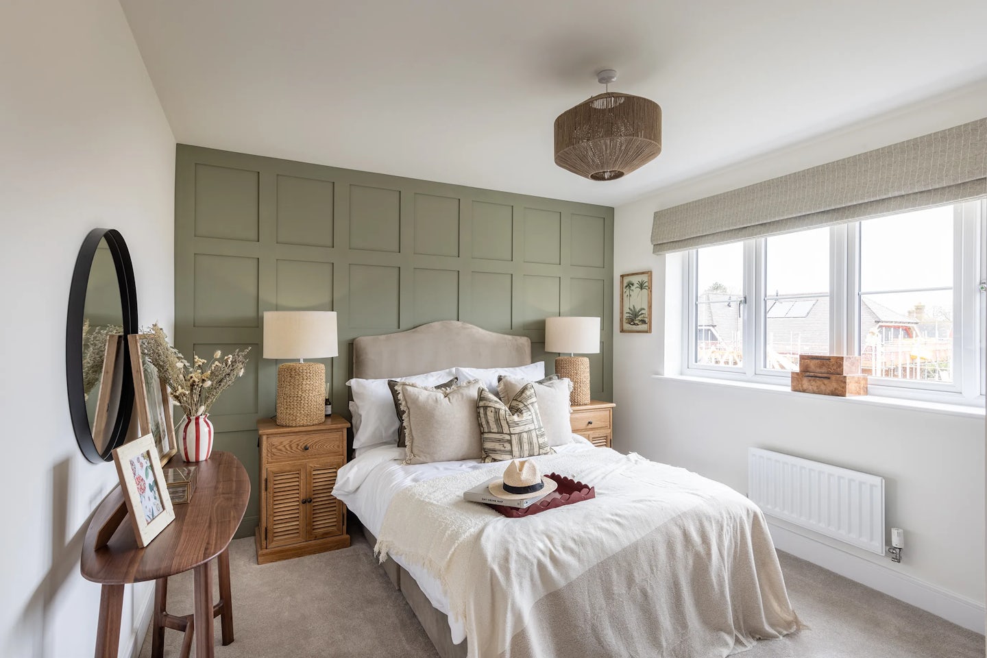 A stylish bedroom with a calming, modern design. A bed with white bedding and layered pillows sits against a green paneled feature wall, flanked by wooden nightstands and soft-glow lamps. A woven ceiling light adds warmth overhead. To the left, a round mirror and console table with decorative accents—such as a vase of flowers and framed art—bring personality to the space. Natural light filters through a large window on the right, above a white radiator, enhancing the inviting and serene atmosphere.