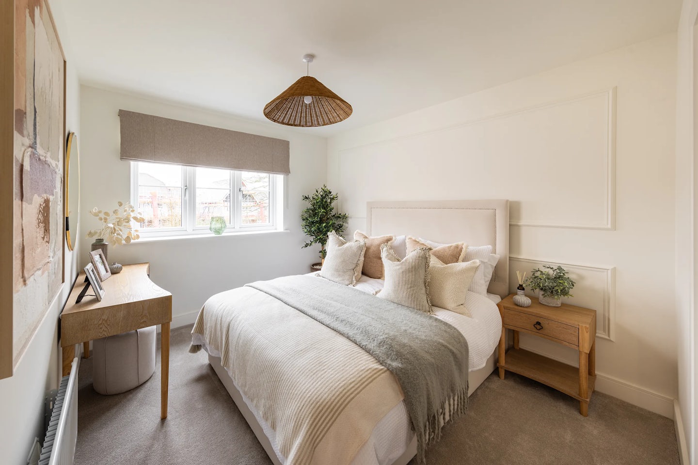 Bedroom with a soft, neutral palette featuring a bed with a beige headboard, layered pillows, and a throw blanket. A wooden nightstand holds a small plant and decor. To the left, a compact wooden desk with decorative items sits beneath abstract wall art. A tall plant is positioned by a window dressed with a beige blind. The room has white walls, carpeted flooring, and a pendant ceiling light. Would you like a version tuned more closely to a specific tone—like more emotive, minimalist, or functionally descriptive?