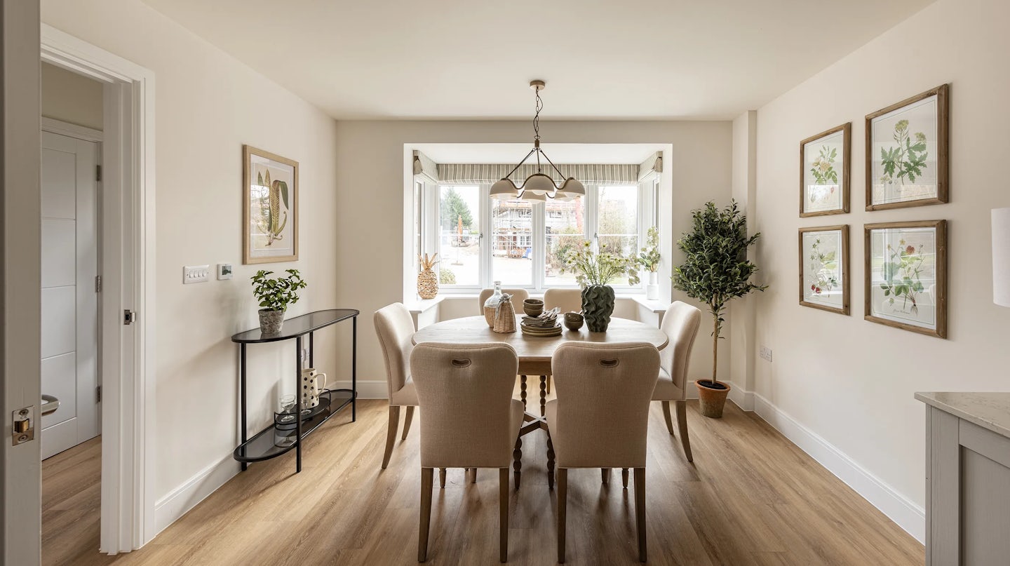 A warm, softly lit dining room featuring a central wooden table surrounded by six beige upholstered chairs. A sculptural vase, woven basket, and decorative centerpiece rest atop the table beneath a classic chandelier. To the left, a console table holds small plants and ornaments, while a bay window with potted greenery allows natural light to pour in. On the right wall, four framed botanical prints add visual interest. The room is styled with neutral tones and organic textures, evoking a cozy, inviting atmosphere.