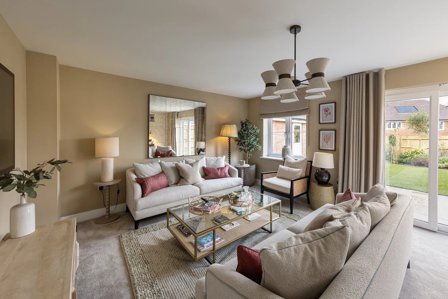 A warm, modern living room with two beige sofas accented by red cushions, a glass coffee table with decor, a large mirror, and a chandelier with multiple lampshades. Additional details include a beige armchair, potted plant, framed artwork, and wide windows with garden views.