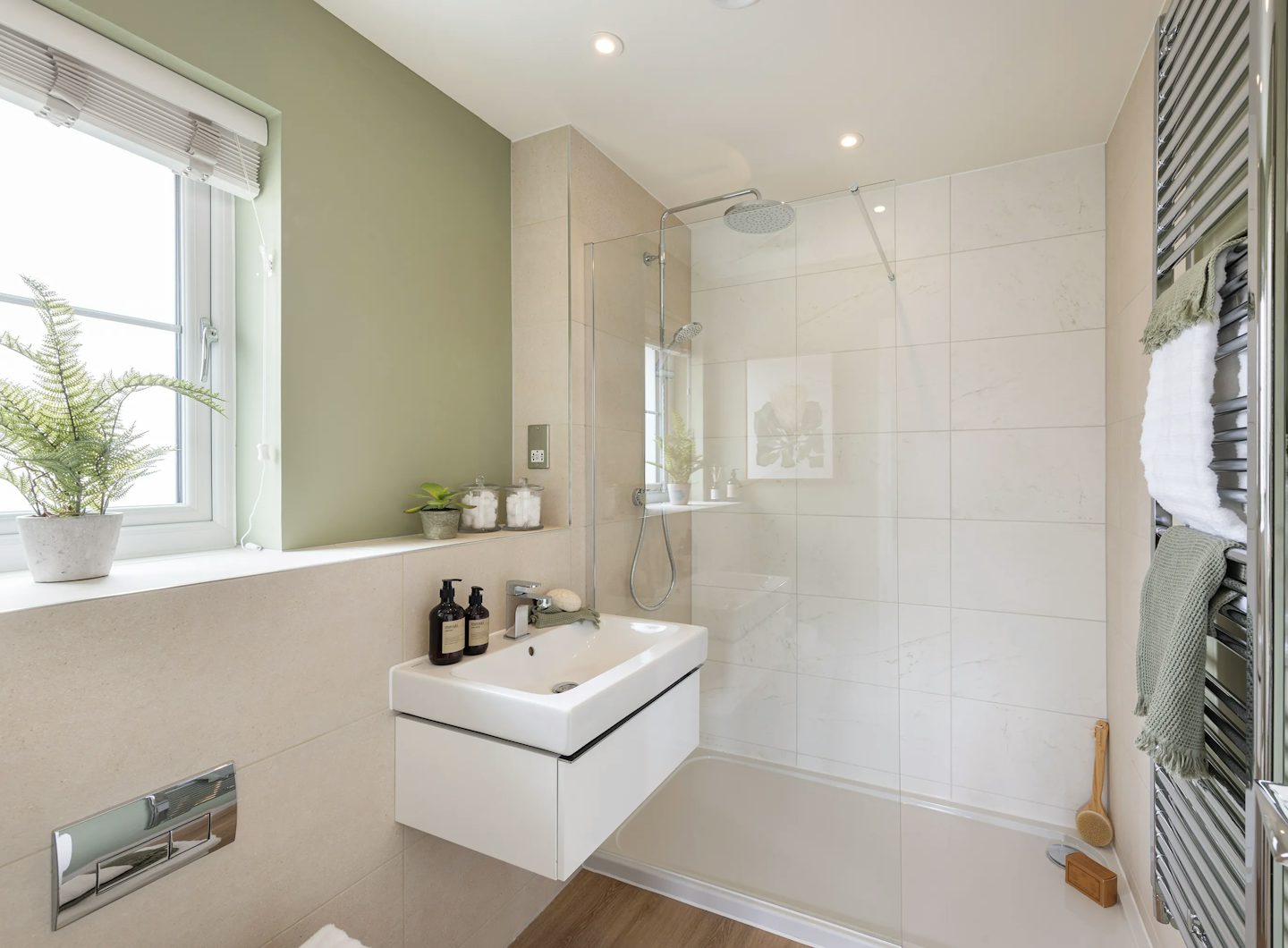 A minimalist bathroom with light green and beige walls, featuring a glass-enclosed shower with a rainfall head, a modern white sink with wall-mounted faucet, and two soap bottles on the counter. A window above the sink is dressed with a white blind and a small potted plant on the sill. To the right, a green towel hangs on a metal rack. The wood-style flooring and recessed ceiling lights add a warm, modern feel.