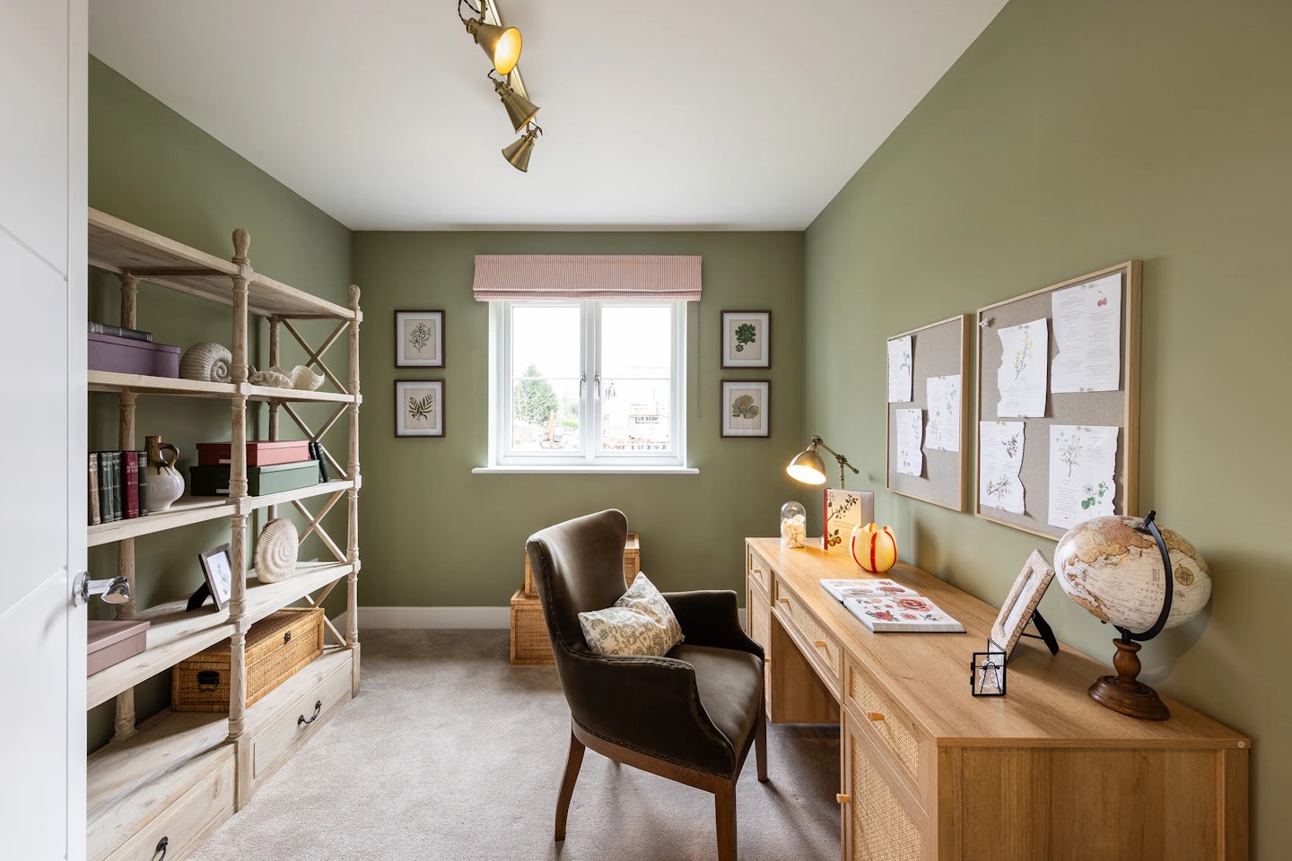 A cozy, organized home office with green walls and a beige carpet. A wooden desk on the right holds a globe, lamp, pumpkin ornament, and scattered papers beneath a corkboard pinned with notes. A wooden shelving unit on the left displays books, storage baskets, and decorative items. A cushioned armchair with a patterned pillow sits centrally, facing a window with a pink blind and two framed botanical prints. Track lights on the ceiling complete the well-lit, welcoming atmosphere.