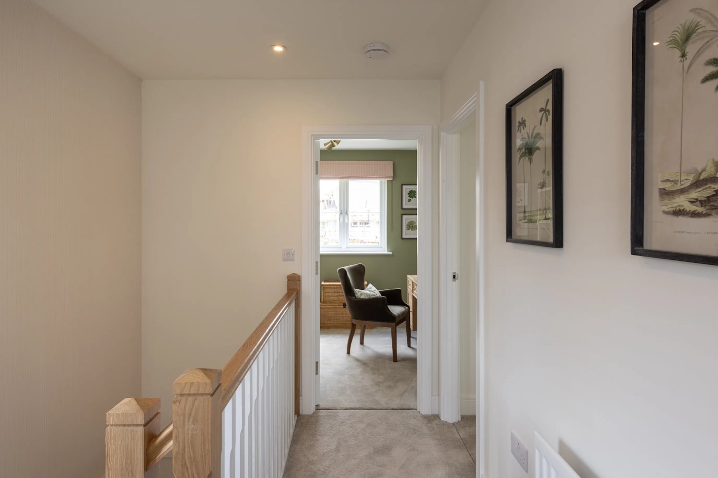 hallway with beige walls and a light carpeted floor leads toward a softly lit room. On the left, a wooden handrail runs along the length of the hall, while framed pictures decorate the right wall. At the end of the corridor, a room with pale green walls features a large circular window that brings in natural light, illuminating a wooden desk and chair beneath it. The space evokes a sense of quiet order and comfort.