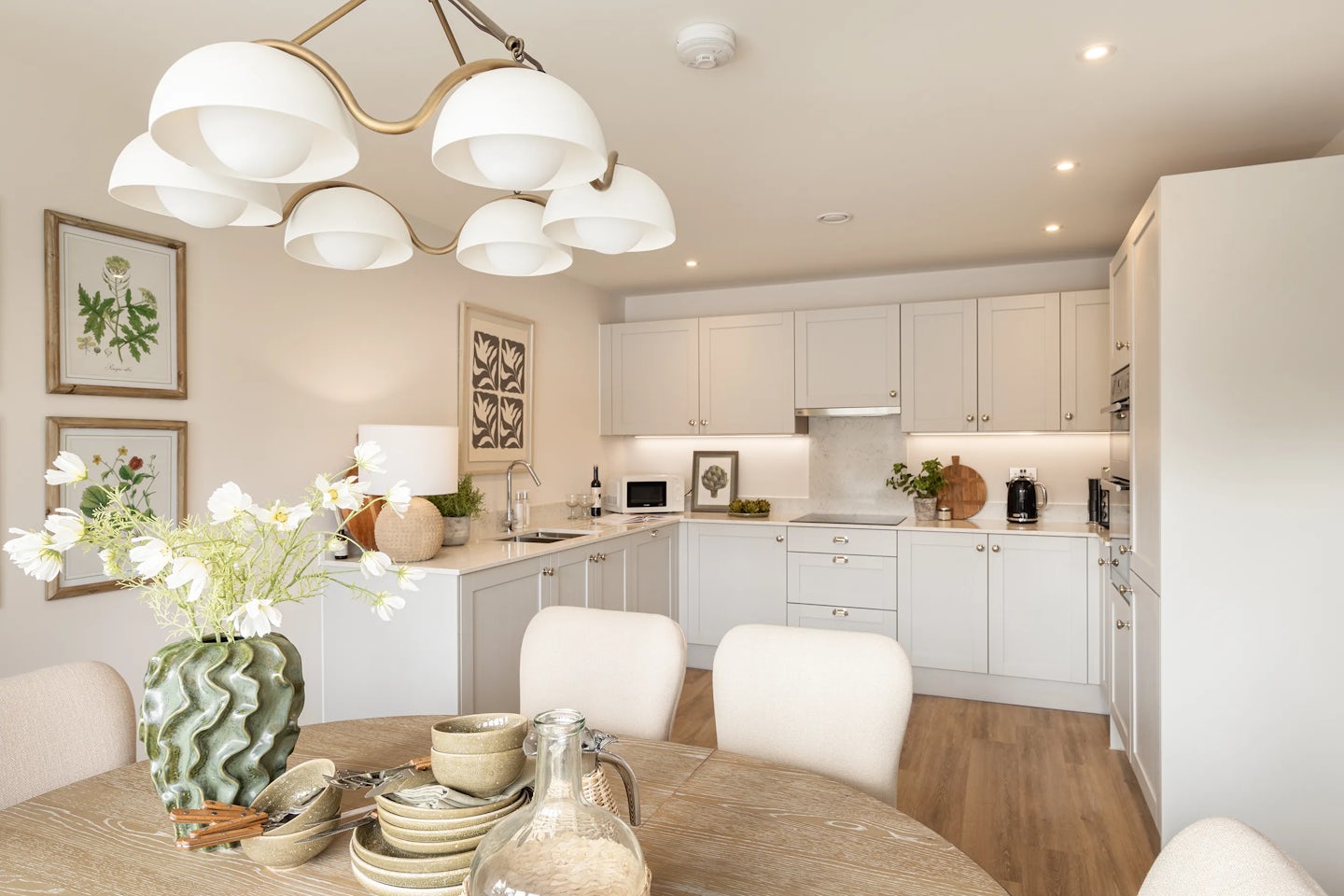 A bright, contemporary kitchen and dining space featuring white cabinets, a white countertop, and stainless steel appliances. A wooden dining table is surrounded by beige upholstered chairs, with a vase of white flowers as a centerpiece. Framed botanical prints hang on the wall above the table. The overhead light fixture has several white lampshades. Kitchenware and dishes are neatly arranged on the table and counters, contributing to a clean, welcoming atmosphere.