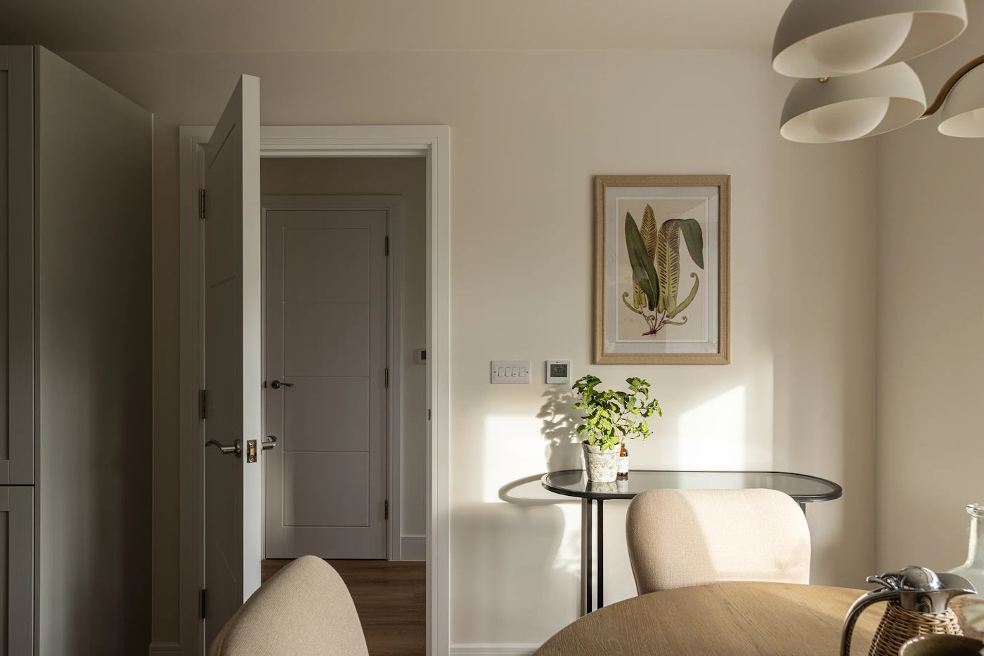 A softly lit interior shows a partially open doorway leading into another room. A botanical print of green leaves hangs above a small round table with a potted plant, adjacent to a light switch and thermostat. In the foreground, a wooden dining table is surrounded by beige chairs, and a modern multi-shade ceiling light adds to the warm, natural ambiance.