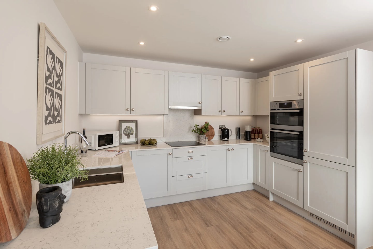 A modern kitchen with white upper and lower cabinets, stainless steel appliances, and a wood floor. A built-in oven and microwave are installed within the cabinetry, and a chrome faucet extends over a stainless steel sink. The countertops hold a mix of everyday items and decorative touches, including a plant, a coffee maker, a cutting board, and small accessories. Recessed ceiling lights provide soft illumination, emphasizing the kitchen's clean, contemporary style.