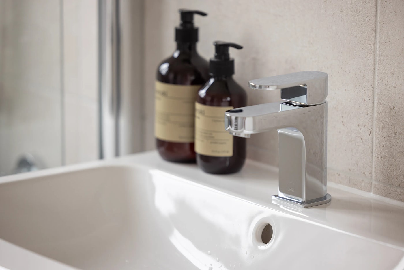 Close-up of a modern bathroom sink with a shiny chrome faucet. Two dark pump bottles with beige labels sit behind the faucet on a white countertop. The sink has a smooth, glossy surface, and the tiled wall in the background adds subtle texture.