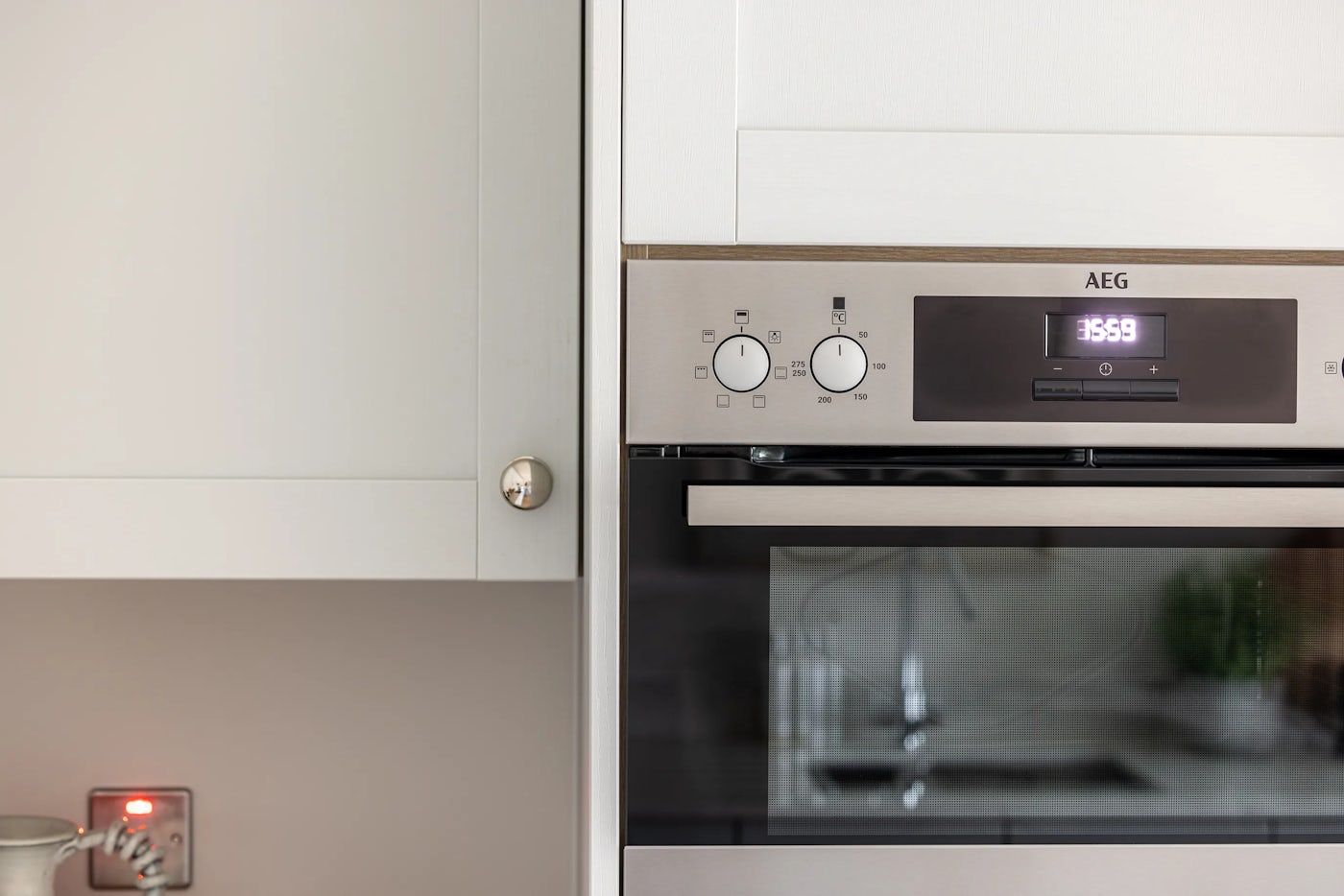 A close-up view of a built-in AEG oven with a reflective glass door. The digital display reads "13:59," flanked by two control knobs—one for oven functions with various cooking mode icons, and the other for temperature settings marked from 50 to 250 degrees Celsius. A faint reflection of a kitchen sink and outdoor greenery is visible in the oven door glass. The unit is integrated into a white cabinet with a pale countertop below.