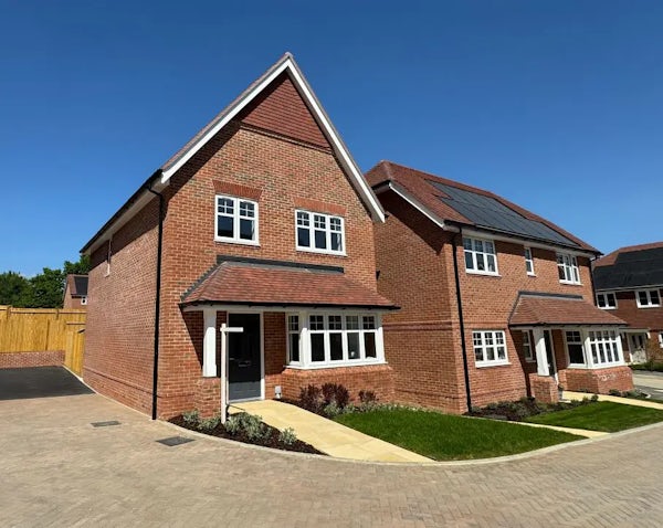 Row of modern brick houses beneath a clear blue sky. The homes feature white-framed windows, black front doors, and pitched roofs. The house on the left includes a garage with a paved driveway, while the house on the right displays solar panels on its roof. Both properties are fronted by neat lawns and simple landscaping along a quiet residential street.